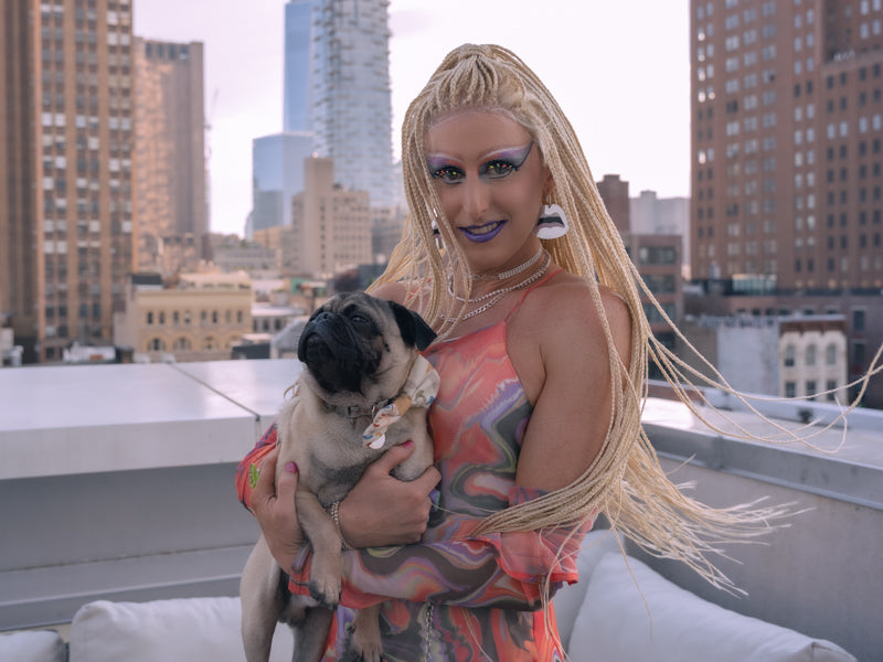 A confident queen in a colorful dress with long, braided blonde hair stands with poised little pug in a bowtie. She is wearing a pair of Bflare Beauty sparkle lashes.