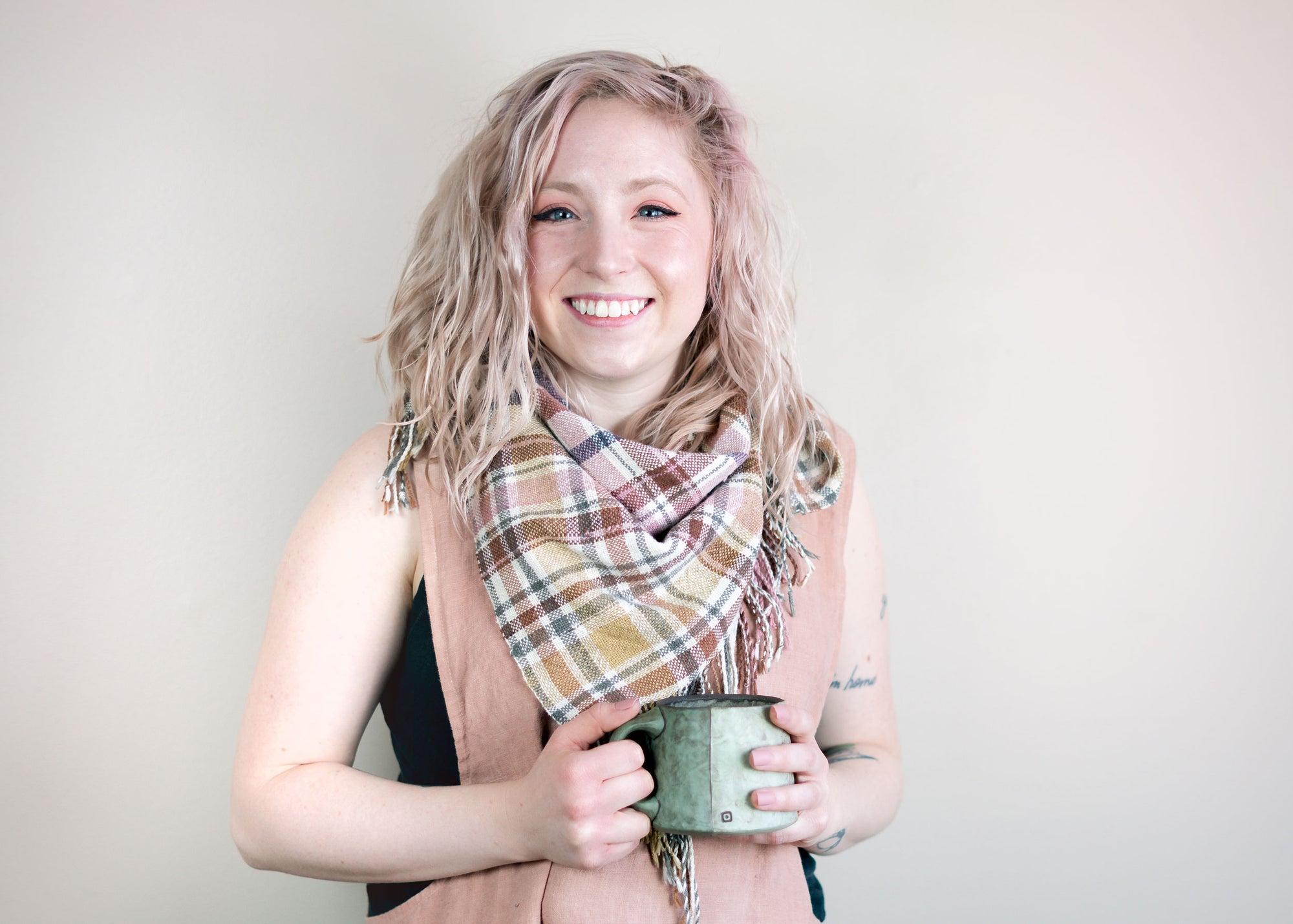 Creative strategist and web designer Emily Harrison of EHarrison Studio smiles at the camera while holding a coffee mug for dear life. She's wearing a handmade apron and scarf. Her hair is wavy and light pink, matching the colors in her scarf. 