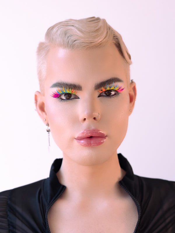 Sean, pictured from the shoulders up, faces the camera straight on with his gorgeous face and perfectly swooped blonde hair. He's wearing a pair of rainbow eyelashes that glow in UV light.
