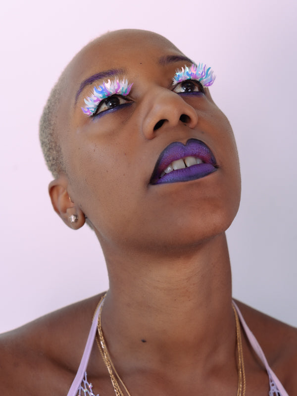 Certified badass Noele looks up away from the camera, wearing a metallic purple lip color that pairs perfectly with white and purple holographic flame lashes by BFlare Beauty.