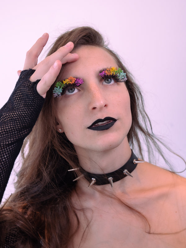Ava looks just above the camera, showing off her rainbow Bloom lashes by BFlare Beauty. She's sweeping some of her long brown hair from her face with a fingerless fishnet gloved hand. 