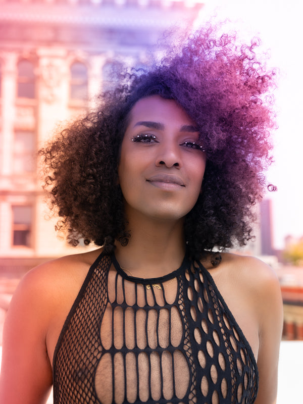 A proud, confident model in a fishnet-style top faces the camera, their curly hair blowing in the wind. They're wearing a simple, custom black and white sparkle Bflare Beauty Lash and the whole scene is cast in rainbow light.
